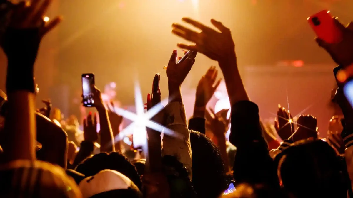 Image of people at an event raising their hands and one taking video on a cell phone.