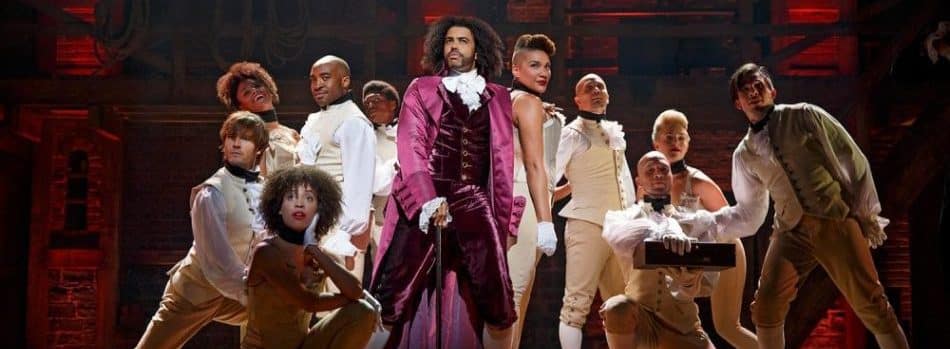 Hamilton cast on stage, surrounding actor Daveed Diggs in costume as Thomas Jefferson